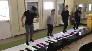 GRUPO TOCANDO PIANO COM OS PÉS , EM PÉ , EM UM PIANO GIGANTE .