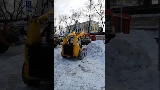 Минипогрузчик и трактор чистят снег и загружают снег в грузовик-самосвал.