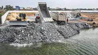 Huge Land Filling Up Reclamation Process By Bulldozer Pushing Rock In Water &Dump Truck Moving Stone